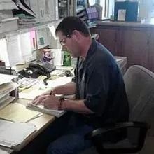 Man seated at a desk typing on a laptop in a cluttered office