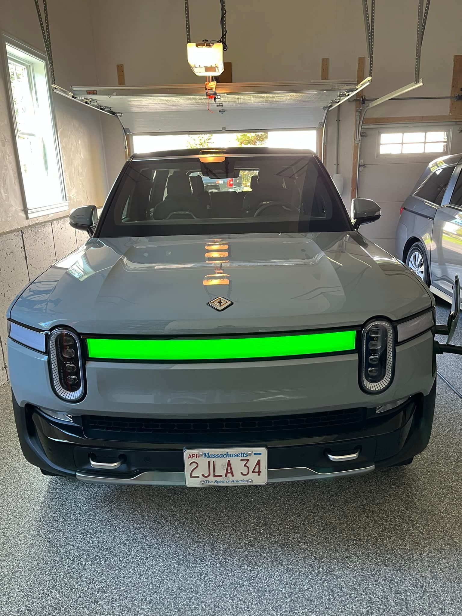 Gray Rivian electric vehicle parked in a garage with green horizontal light bar, Massachusetts license plate.