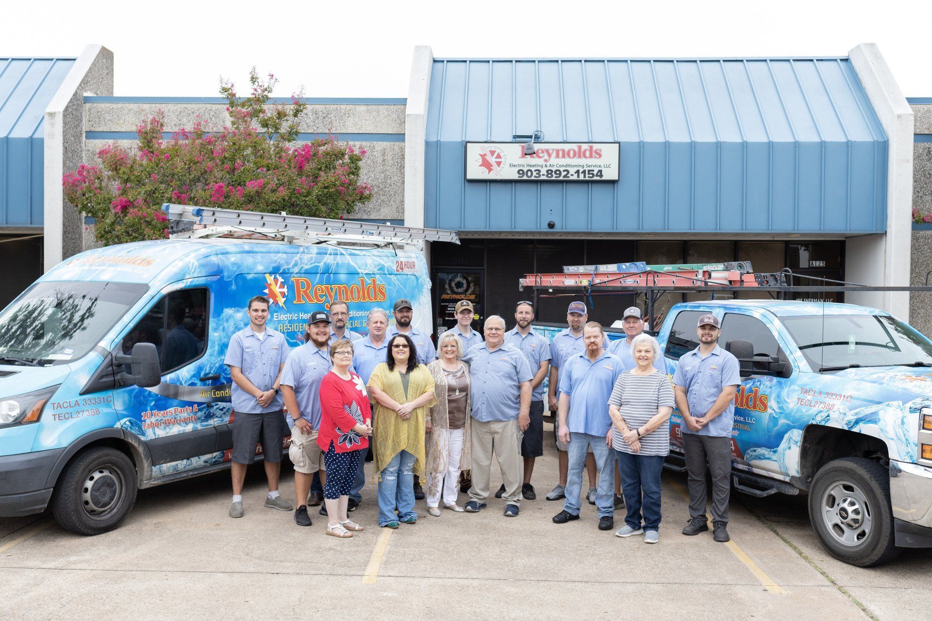 A group of people posing in front of a blue building with two service vehicles. Some wear blue shirts.