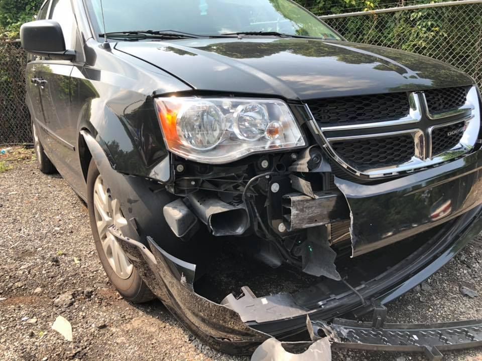 Black minivan with severe front-left damage and missing bumper, parked on gravel.