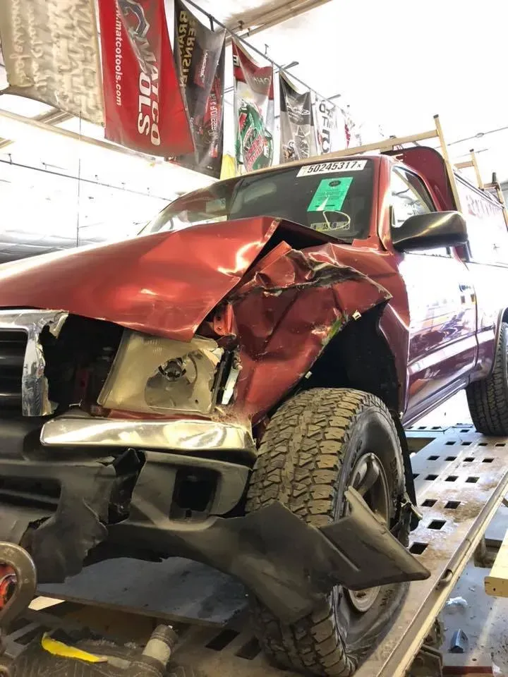 Severely damaged red pickup truck on a shop lift, with crushed front end and front wheel turned outward