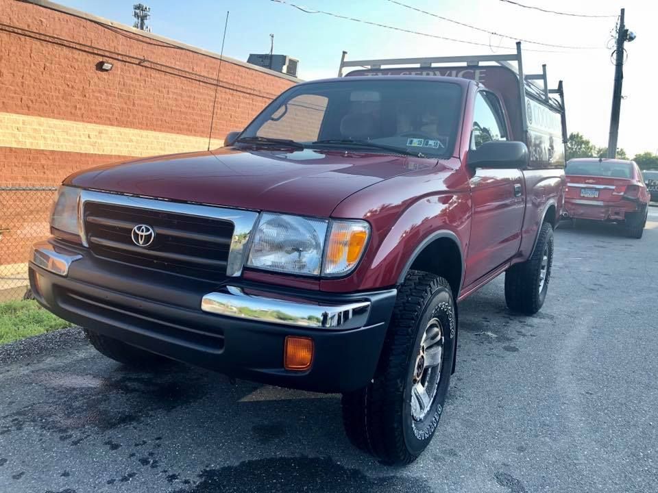 Maroon Toyota pickup truck parked on a street beside a brick building, with another car behind it.