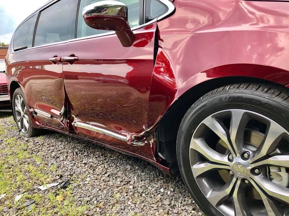 Damaged maroon SUV with crumpled side panel and broken door, parked on gravel.