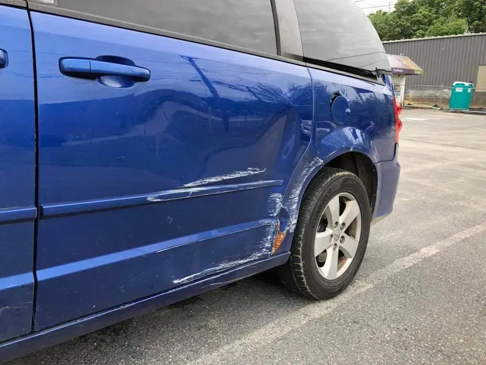 Blue car with side and rear damage, including scratches and dents, parked in a lot.