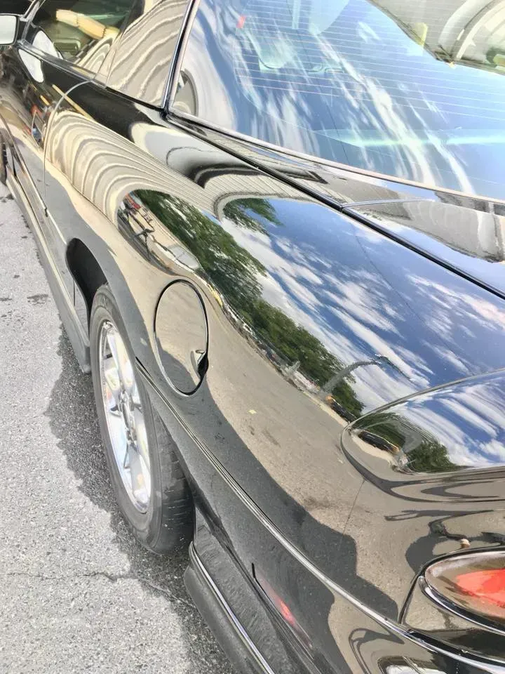 Black sports car close-up, showing rear side, fuel cap, wheel, and sky reflections on the glossy paint.