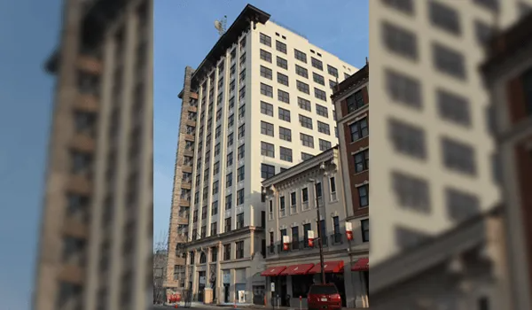 Tall, light-colored building in downtown area with square windows and smaller adjacent buildings.