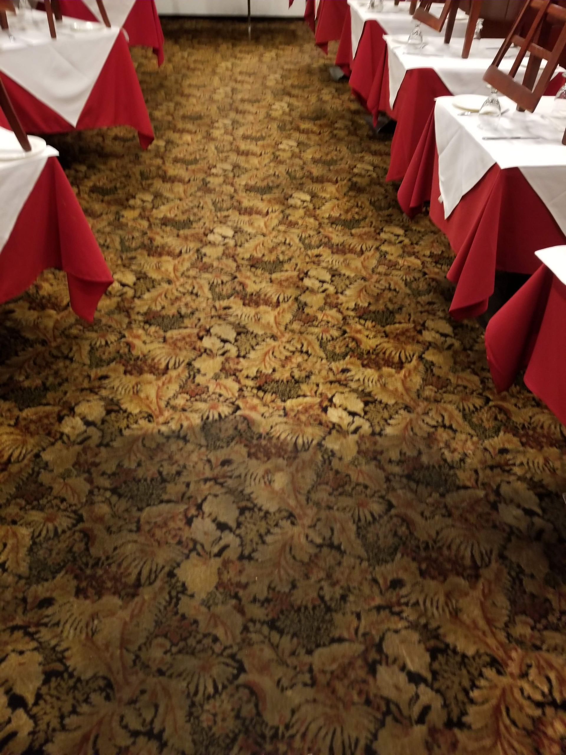 A restaurant dining area with tables set in white tablecloths and red table skirts along a patterned carpeted aisle.