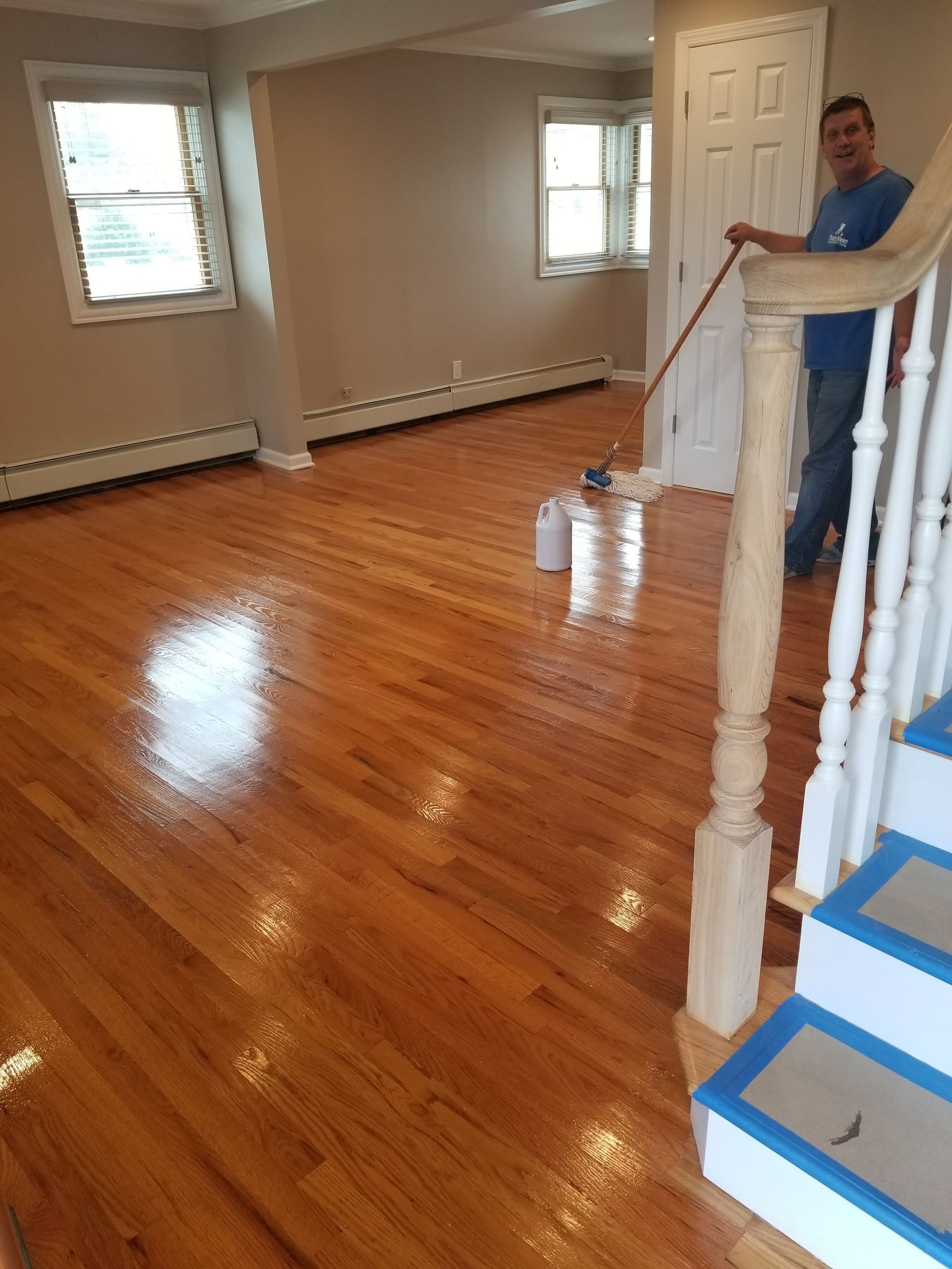 Empty room with polished hardwood floors; a man stands by a staircase railing holding a bucket and mop.