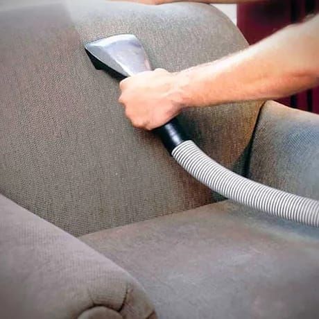 A hand cleaning a gray upholstered chair with a vacuum upholstery attachment