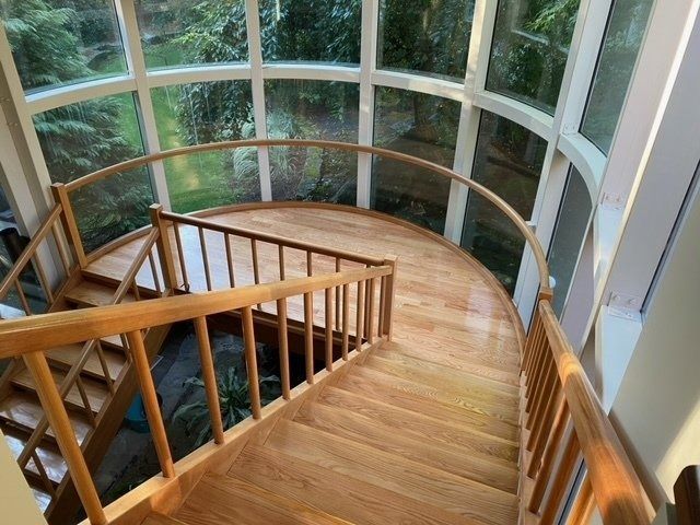 Wooden spiral staircase leading up, enclosed by curved windows, bathed in sunlight.
