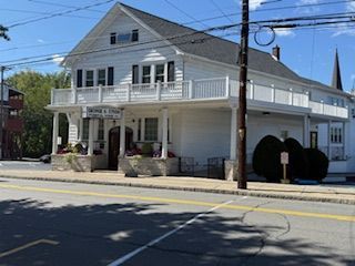 White two-story building with porch and balcony, flowers in front. Sign above door. Sunny day, exterior shot.
