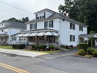 White two-story building with black shutters, awning, and small porch. Driveway and street in front.