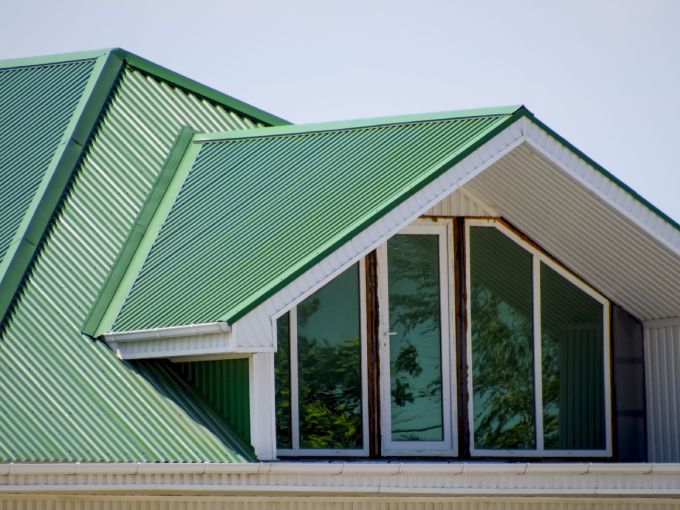 Learn More About Metal Roofing Installation A close-up of a green, corrugated metal roof featuring a gabled dormer with large glass windows and white trim.