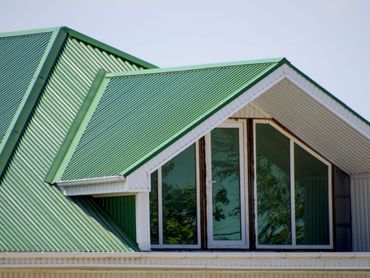 A close-up of a green, corrugated metal roof featuring a gabled dormer with large glass windows and white trim.