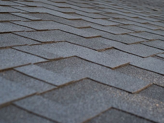 Learn More About Asphalt Shingle Roofing Close-up of gray asphalt roof shingles with overlapping rows and slight weathering