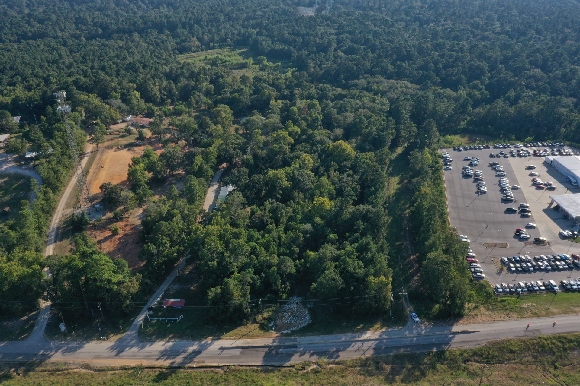 Aerial view of a forested area beside a large parking lot and road, with cars lined up.