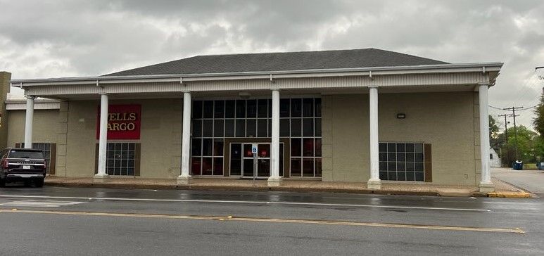 Front view of a vacant storefront with white columns, large windows, and a red sign by a wet street.