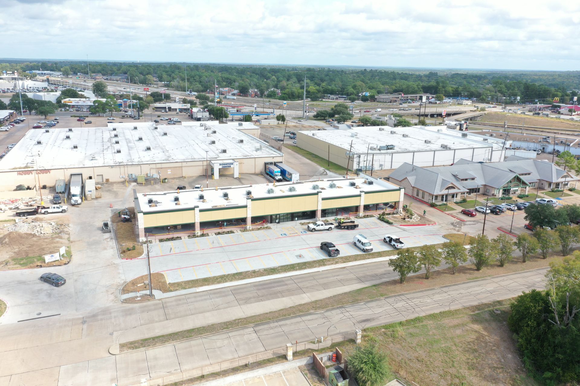 Aerial view of a low-rise shopping complex with parking lots, roads, and surrounding buildings.
