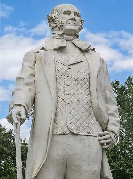 White marble statue of a standing man in a coat and vest, against a blue sky and trees