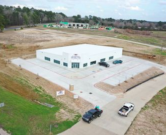 Aerial view of a white industrial building with paved lot, trucks, and open land around it.