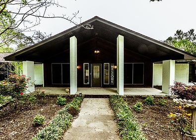 Front view of a small house with a covered porch, central walkway, and surrounding shrubs