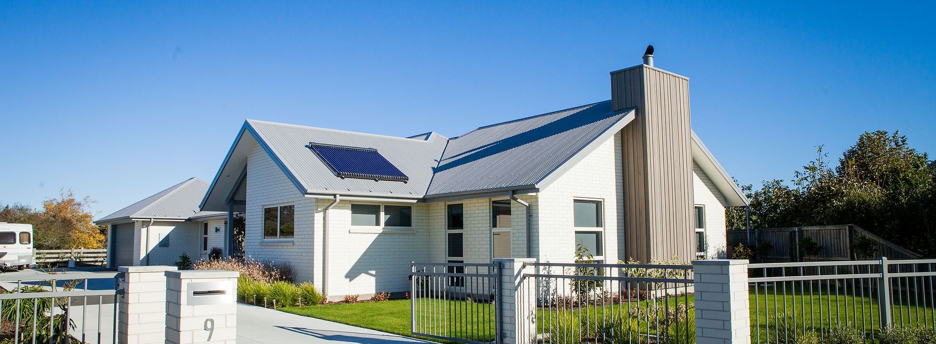 Modern white house with a blue roof and solar panels, behind a black fence on a sunny day