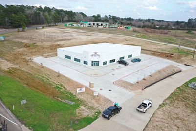 Aerial view of a white industrial building on a concrete lot with two parked vehicles, surrounded by open land.
