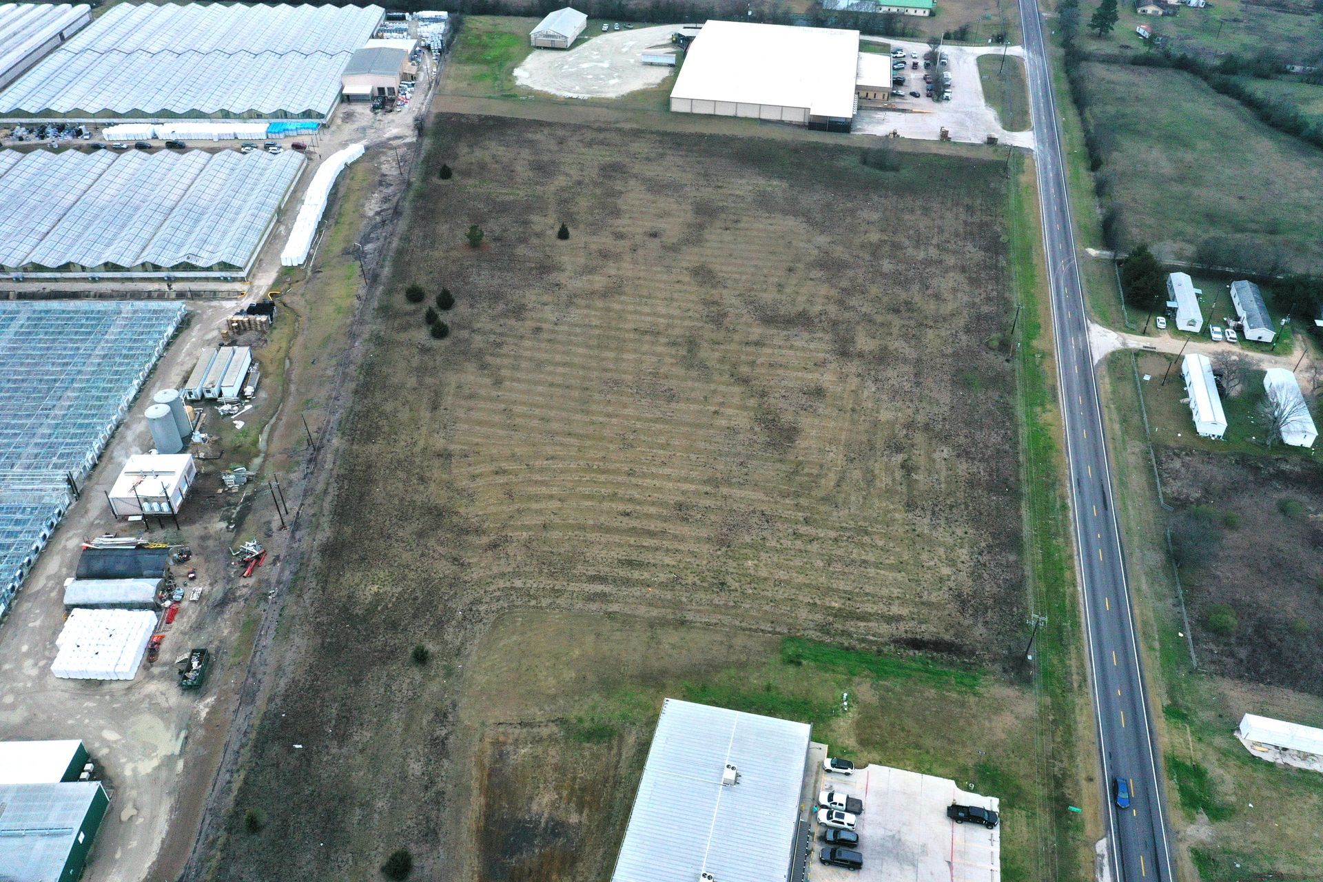 Aerial view of a large brown field beside greenhouses and a road in a rural area
