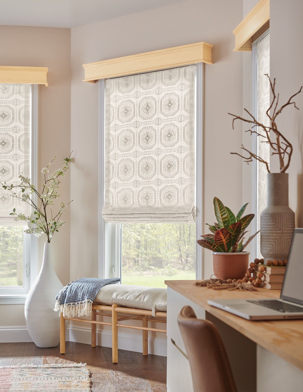 A living room with a desk , chair , vases and a window with roman shades.