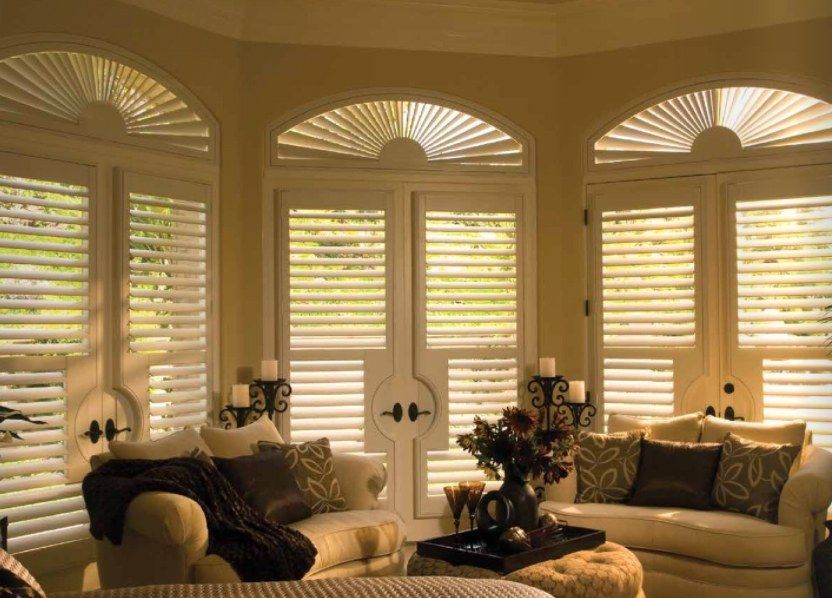 A living room with a couch and ottoman and shutters on the windows
