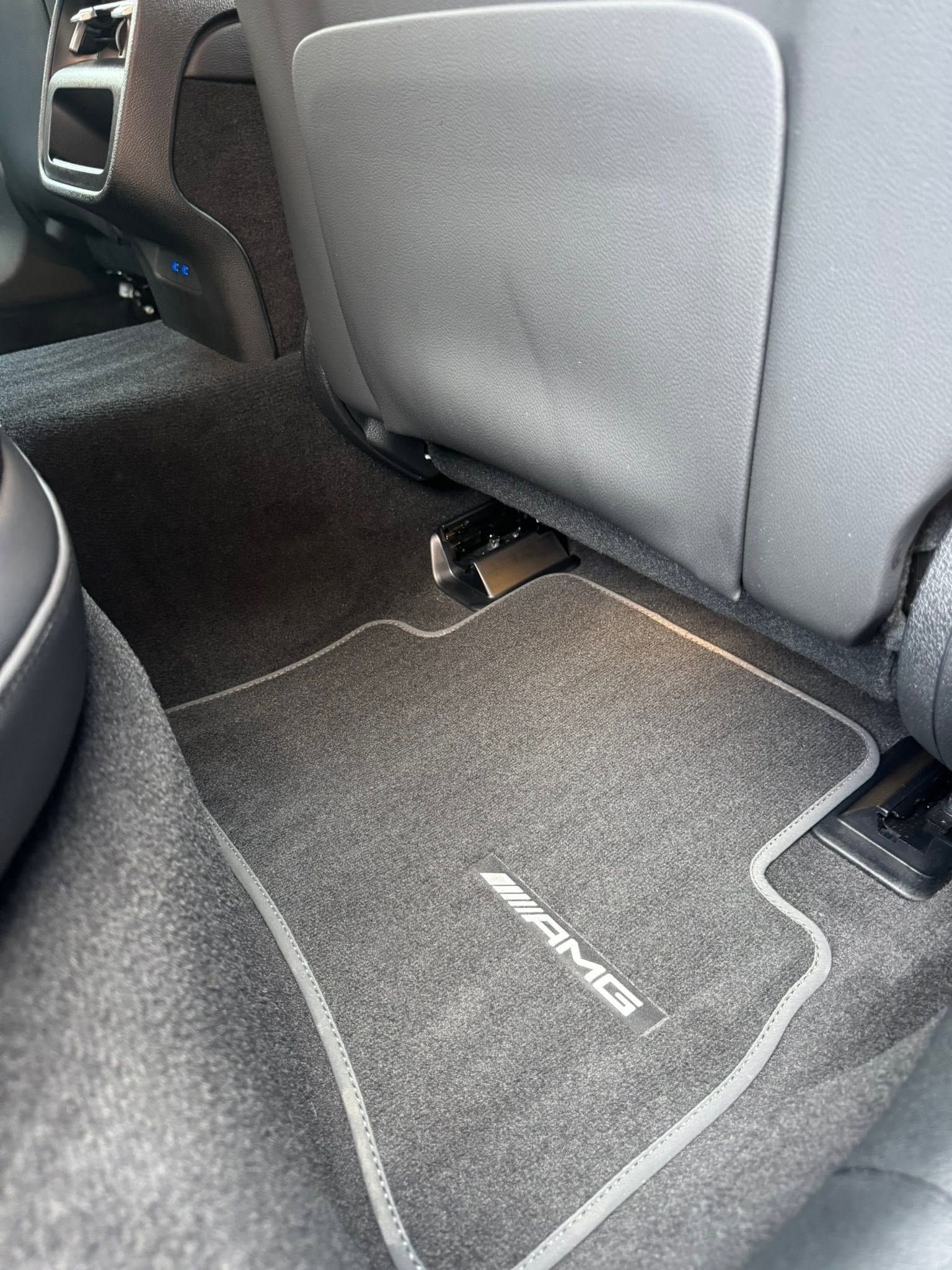 Car floor mat in a vehicle’s rear seat area, dark gray with white trim and logo.