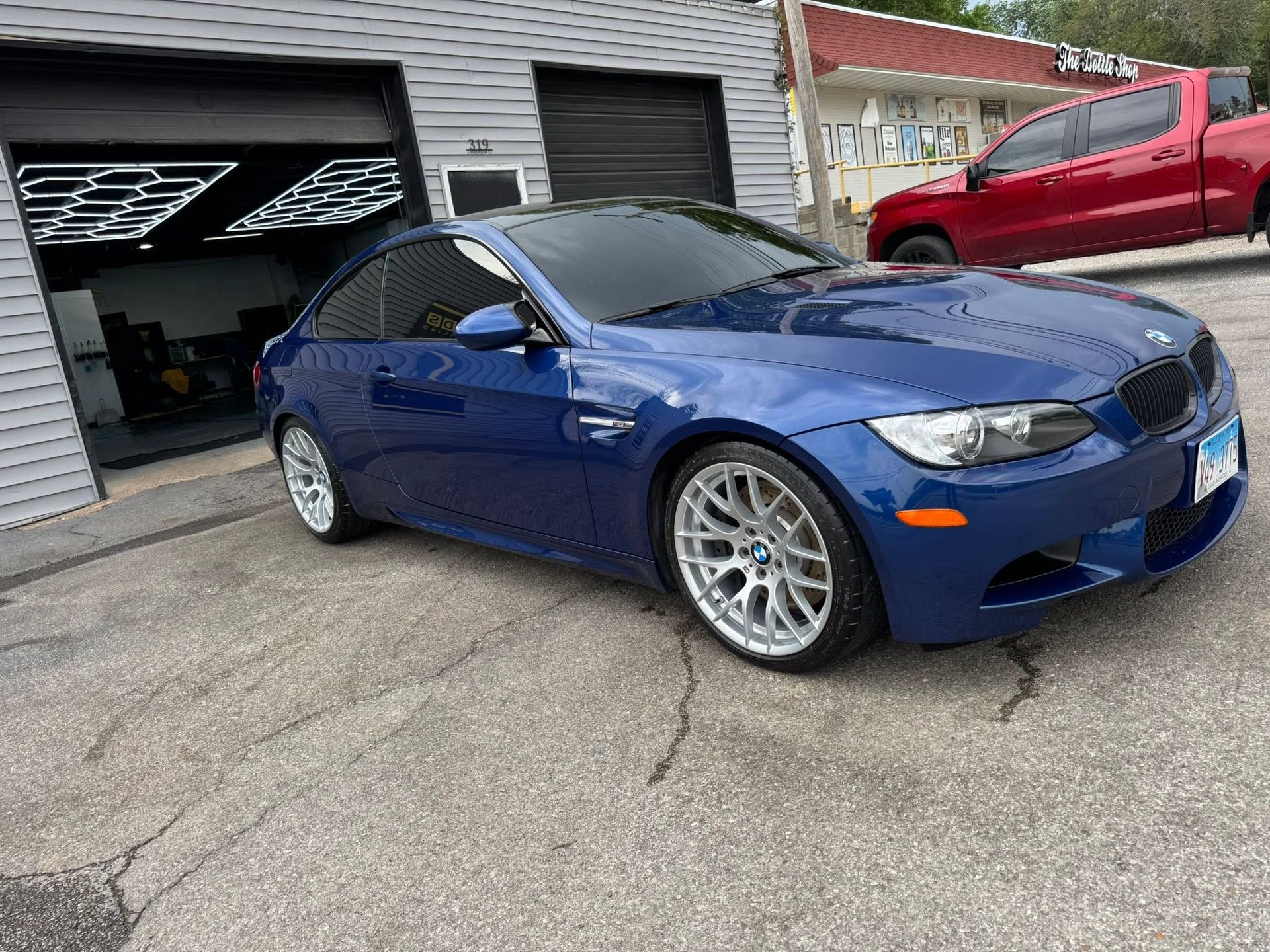 Blue sports sedan parked outside a garage, with silver wheels and a red truck in the background