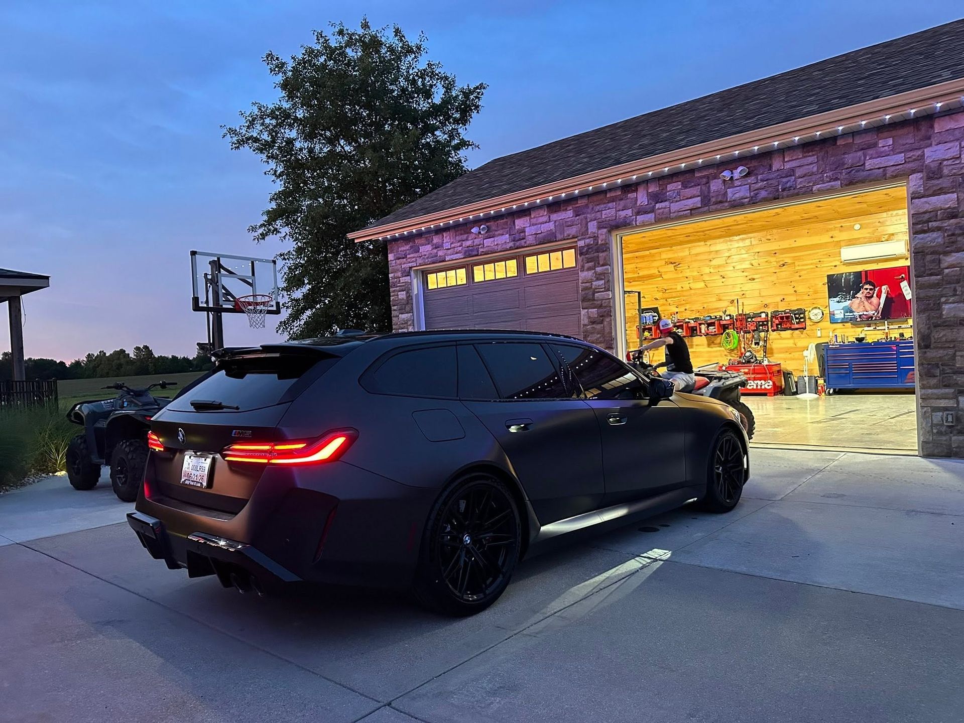 Black sedan parked outside a lit garage at dusk, with another car and a bicycle nearby.
