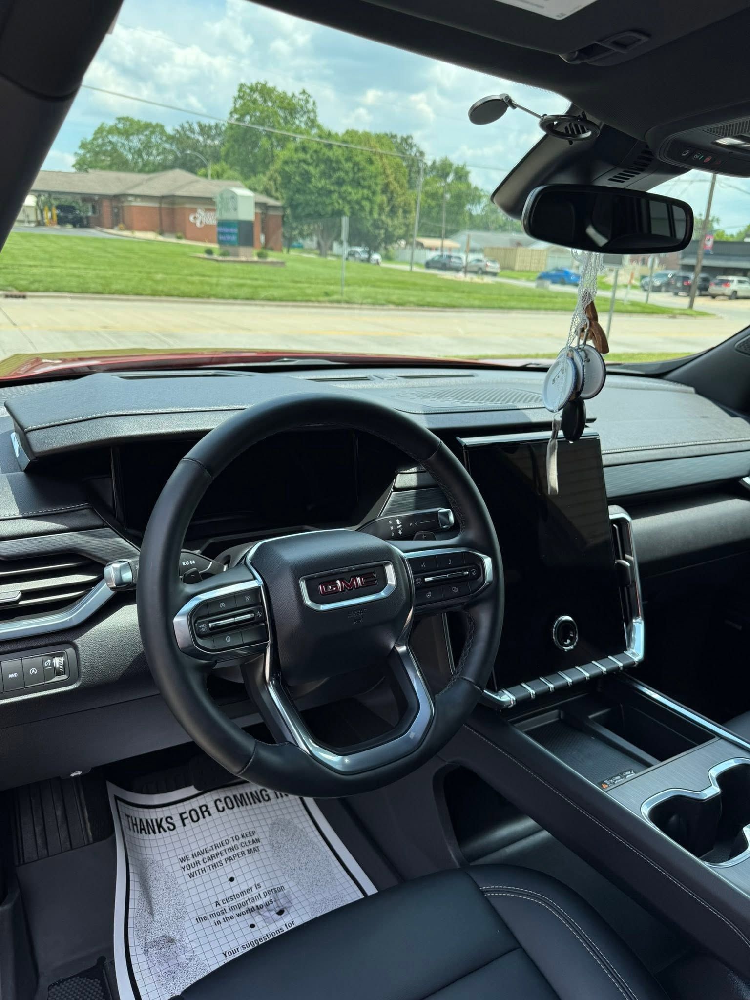 Car interior with steering wheel, dashboard screen, hanging air freshener, and view of a suburban street ahead