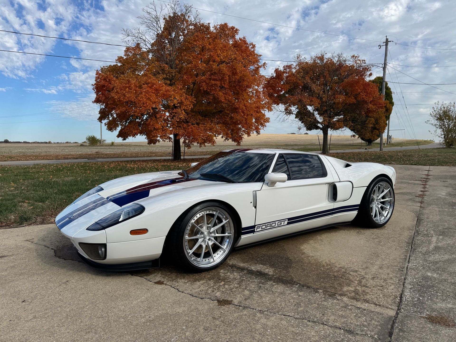 White sports car with blue racing stripes parked on a rural roadside beside autumn trees under a cloudy sky