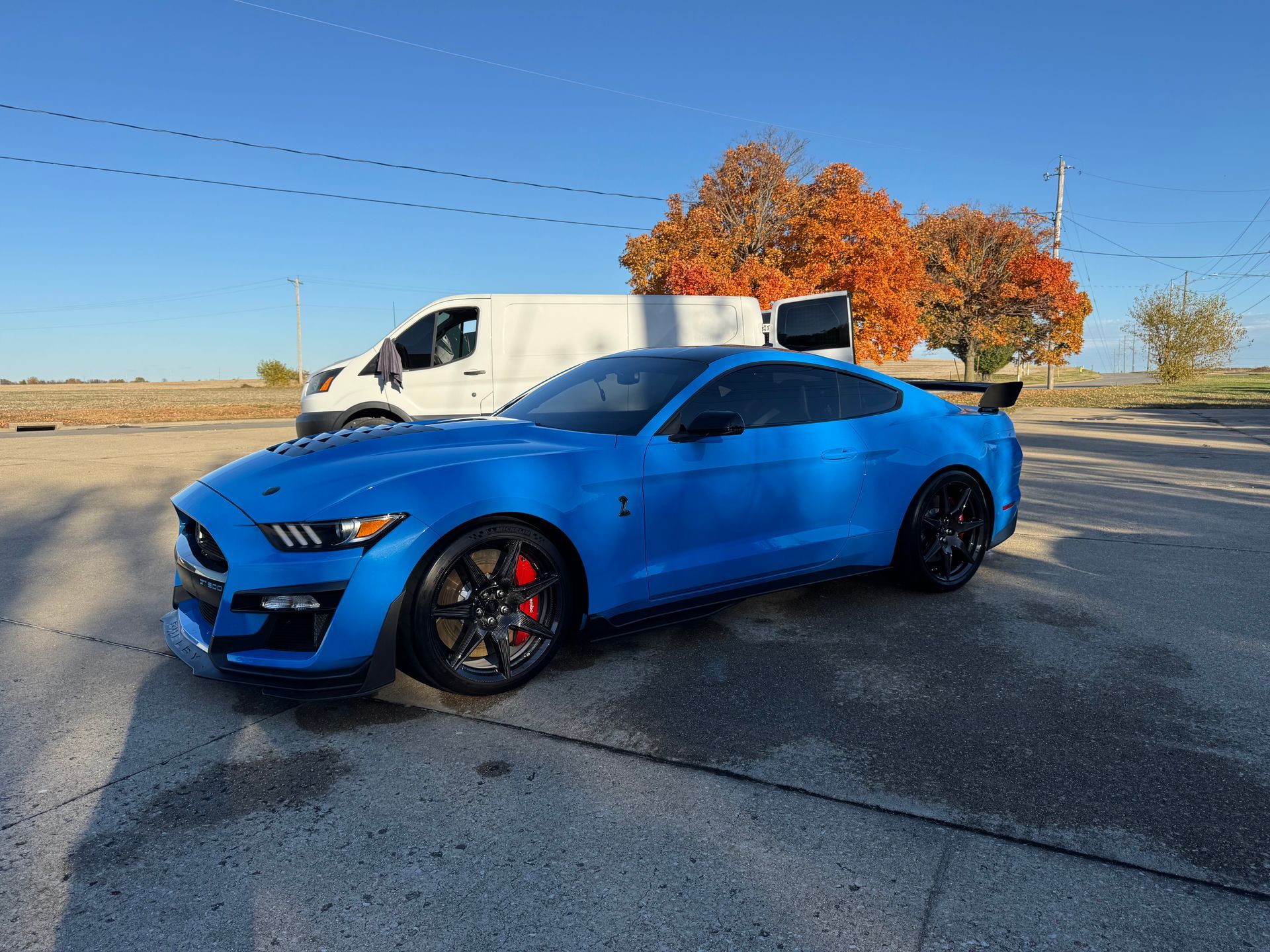 Blue sports car parked on a road beside a white van, with autumn trees in the background.