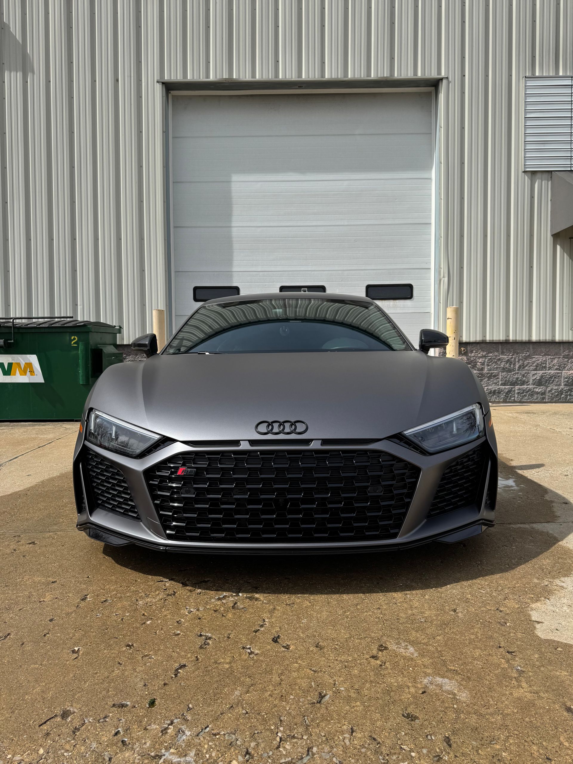 Gray Audi sports car parked in front of a metal garage door on a gravel lot.