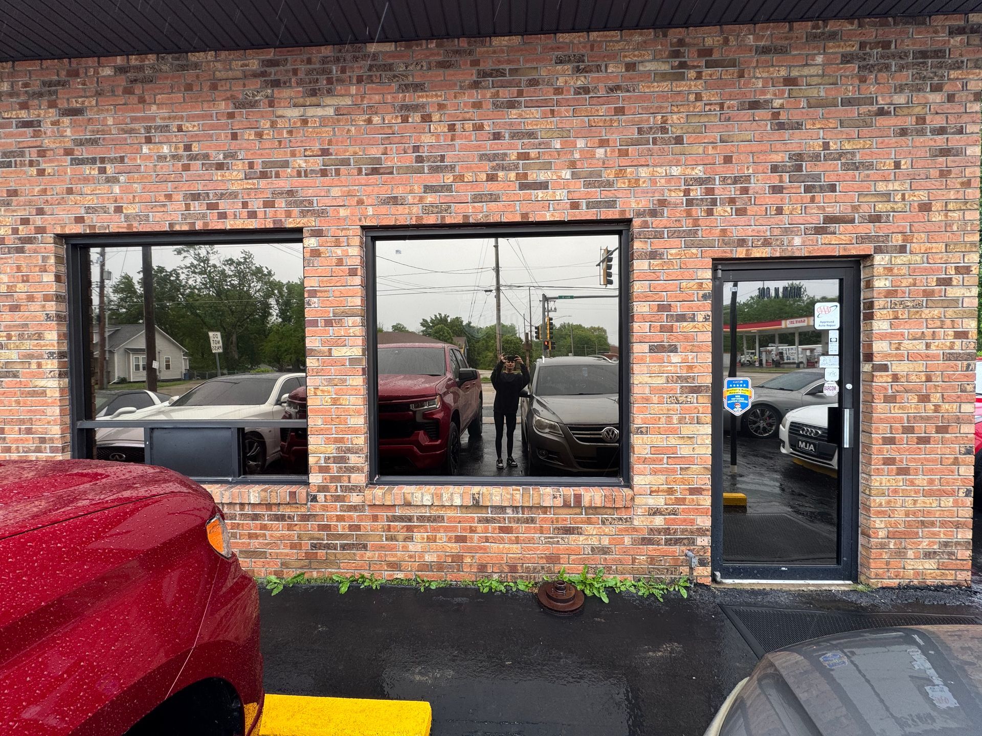 Brick storefront with parked cars reflected in windows and a person standing by the center window.