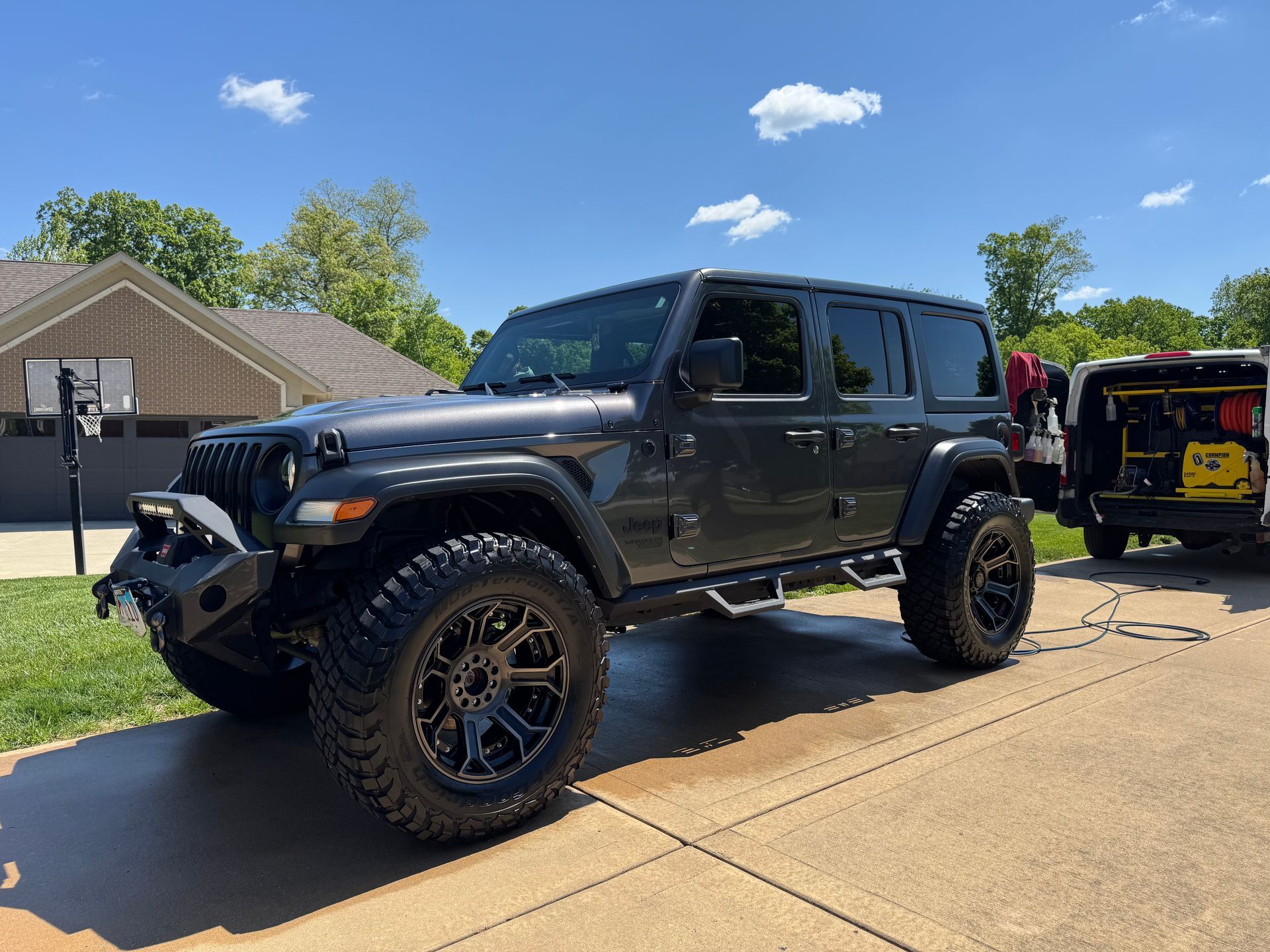Black lifted Jeep Wrangler parked on a sunny driveway beside another off-road vehicle.