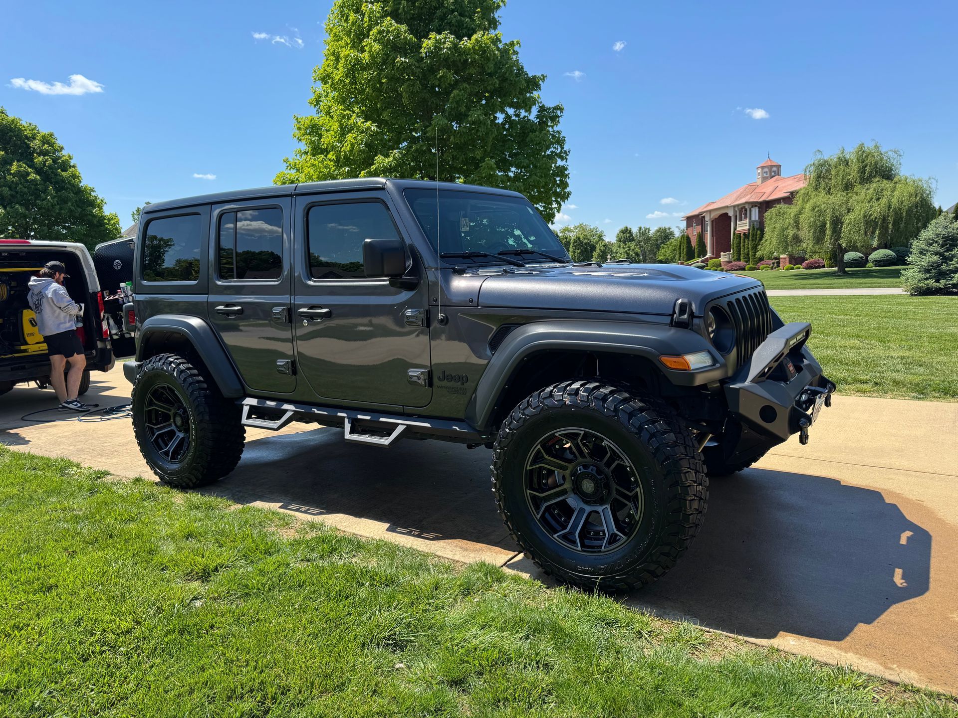 Black Jeep Wrangler parked on a sunny road beside a grassy park, with people in the background