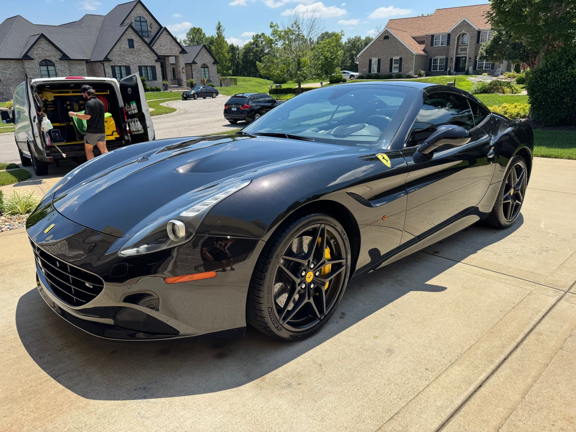 Black Ferrari convertible parked in a suburban driveway, with a garage and houses in the background