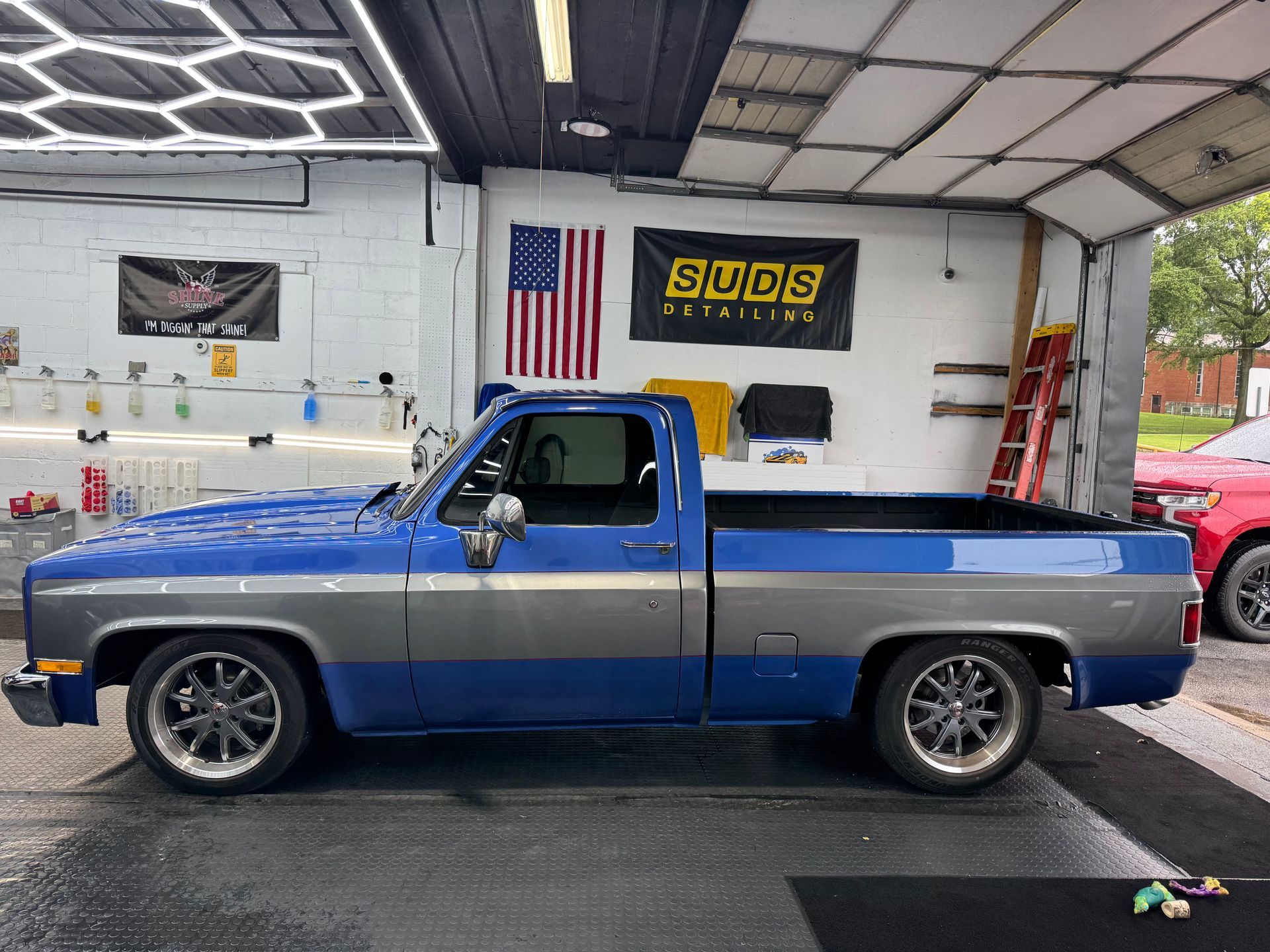 Blue and silver lowered pickup truck in a garage, side view, with an American flag and shop signs behind it
