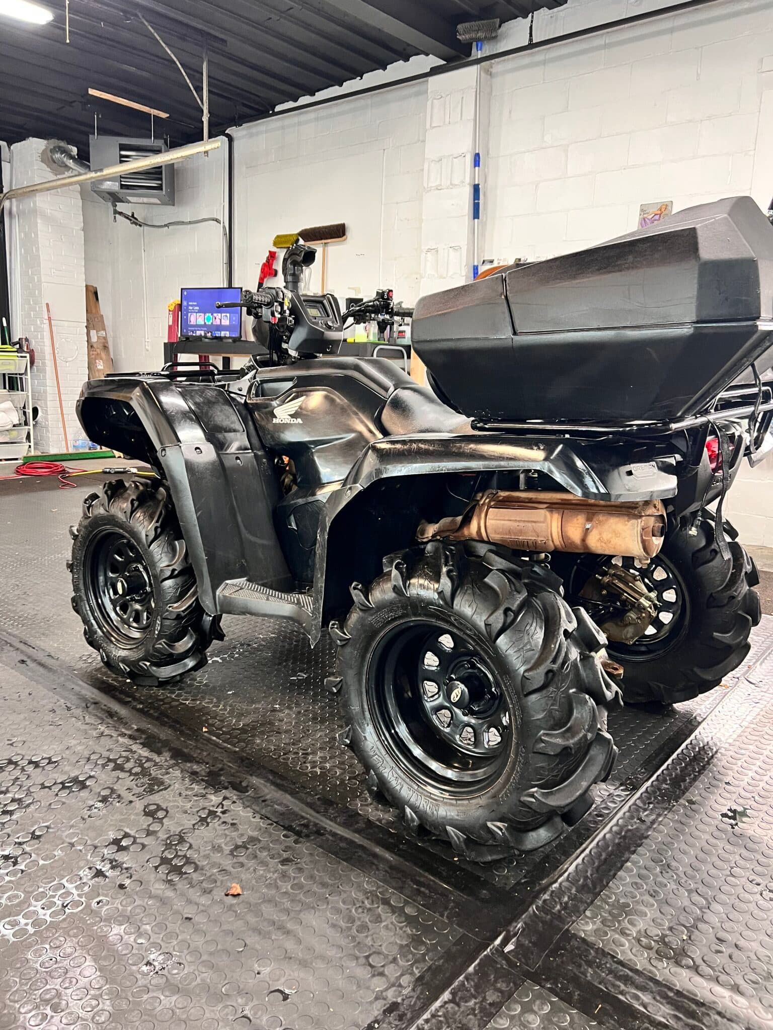 Mud-covered black ATV in a garage, viewed from the rear-left.