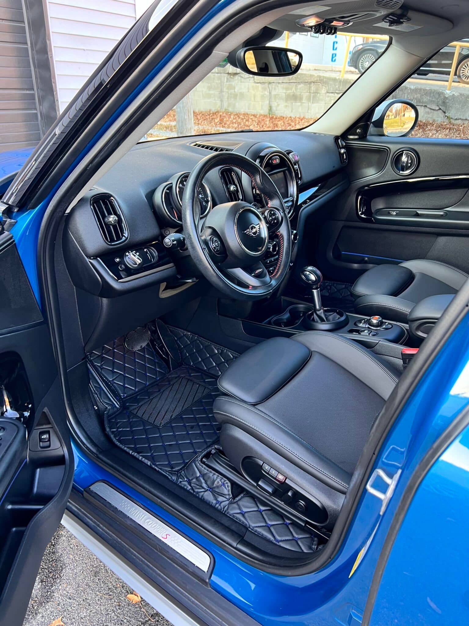 Open driver’s door of blue car, showing black interior and steering wheel with patterned floor mats.