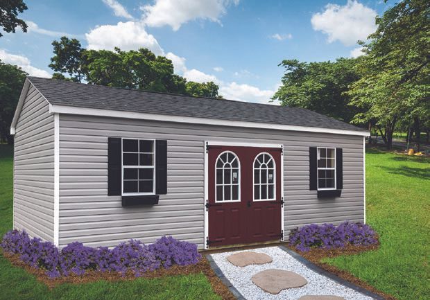 Gray shed with black shutters, burgundy double door, and a stone path. Purple flowers in the front.