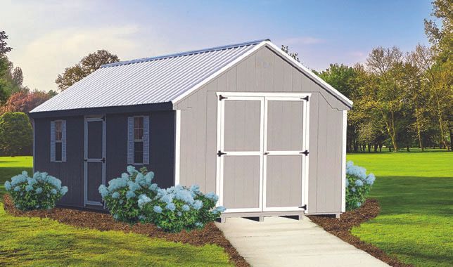 Gray shed with a white-framed double door and windows, surrounded by bushes, on a grassy lawn with a concrete walkway.