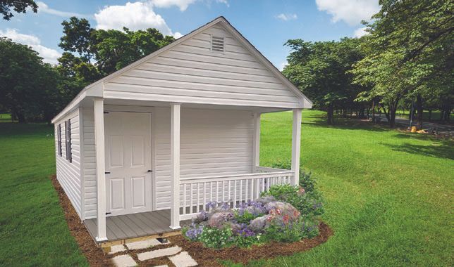 White shed with porch, door, and small garden in front. Green grass and trees surround it.