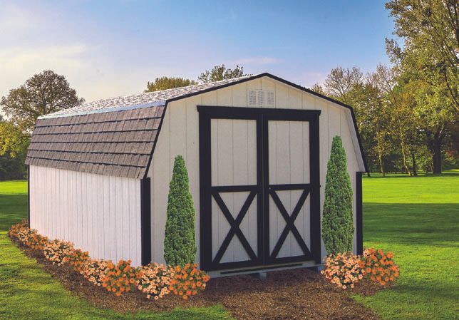 White barn-style shed with black doors, surrounded by landscaping, set in a grassy yard.