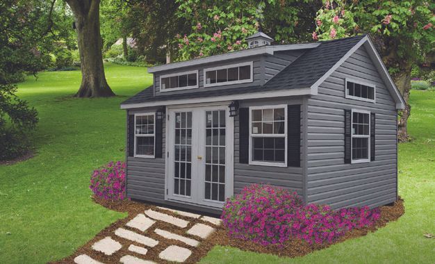 Gray shed with French doors, small dormer windows, shutters, and a stone path in a garden setting.