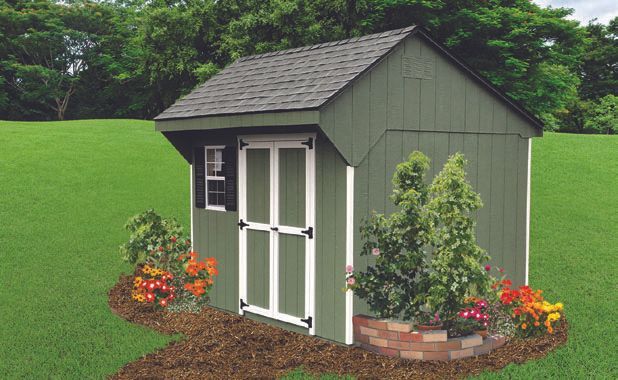 Green wooden shed with white-trimmed doors and a small window, surrounded by flowers and a grassy lawn.